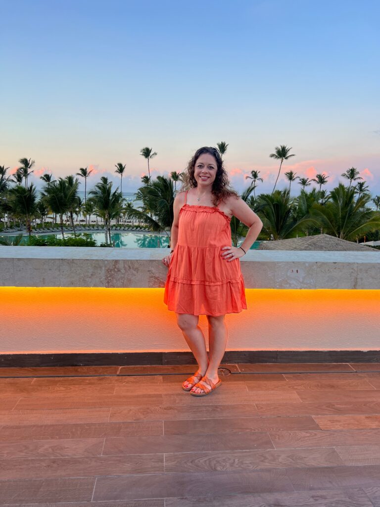 Picture of a woman standing on a rooftop with palm trees, a pool, and a beach in the background.